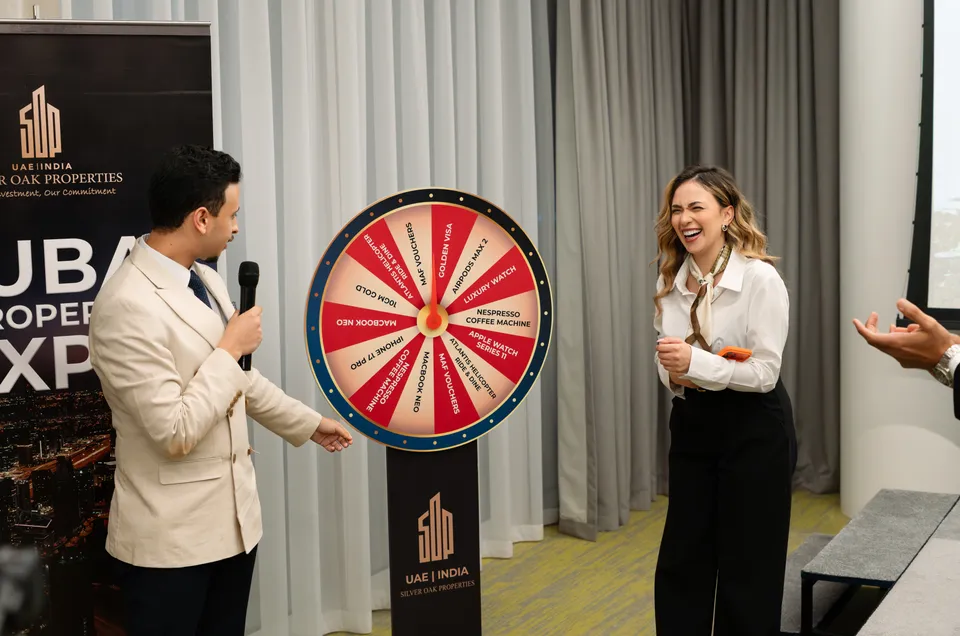 A host with a microphone stands beside the Silver Oak Properties prize wheel as a guest laughs