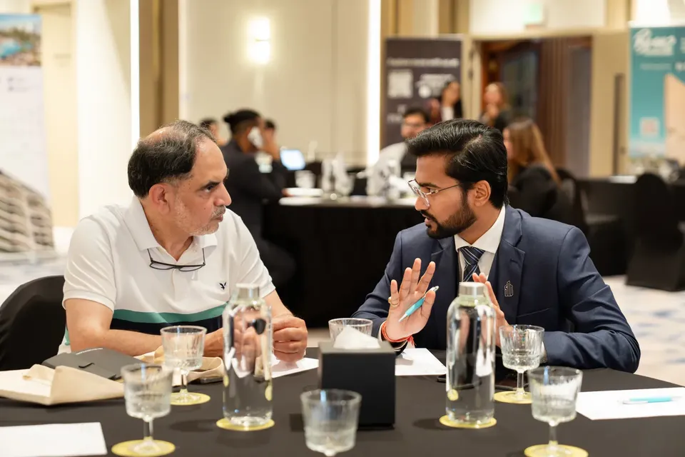 An adviser in a navy suit gestures while speaking with a seated guest in a white polo shirt at a consultation table