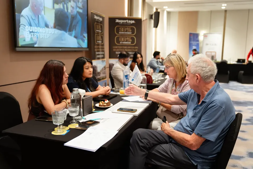 Across the table, two advisers meet with an older couple, beside a Reportage banner and a screen of step-by-step guidance