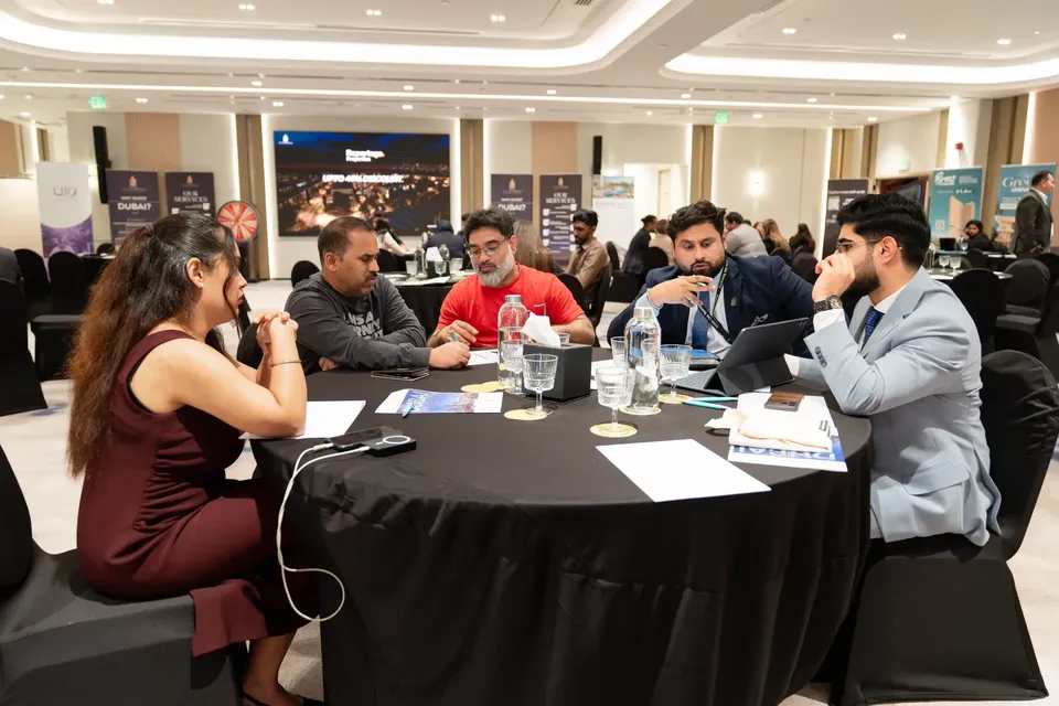 Five people sit in discussion around a round table on the expo floor, with rows of tables beyond