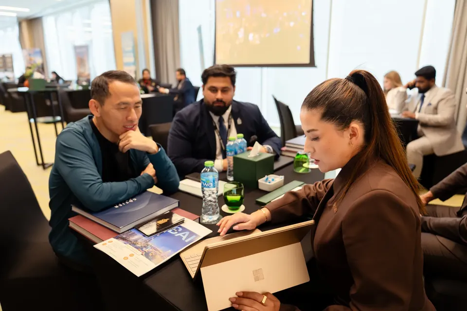 An adviser in a brown blazer shows a tablet to two seated visitors at a consultation table lined with water bottles