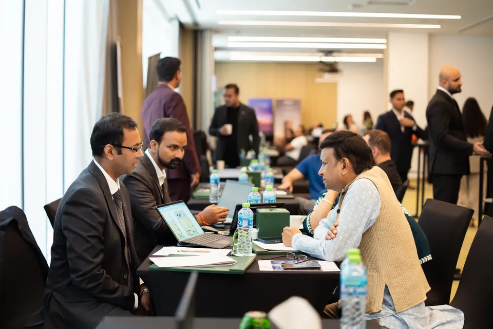 Two suited advisers consult with a visitor in a beige waistcoat over a laptop at a busy expo table