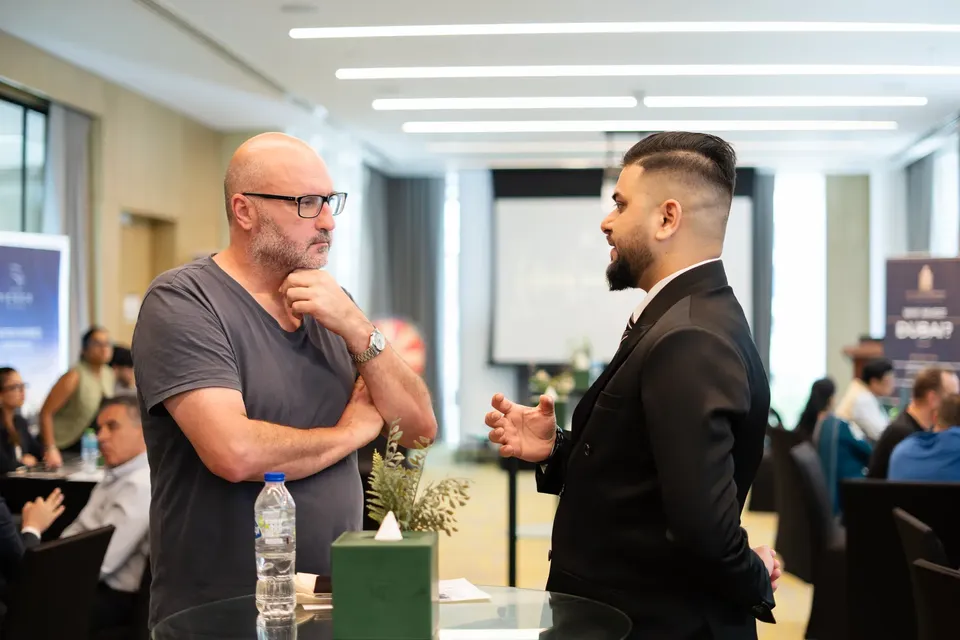 Standing beside a small table, a visitor in a grey t-shirt talks with an adviser in a dark suit
