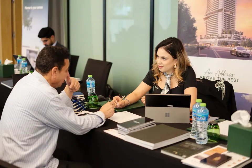 A visitor sits with an adviser at a consultation table, a tower render on the display behind her