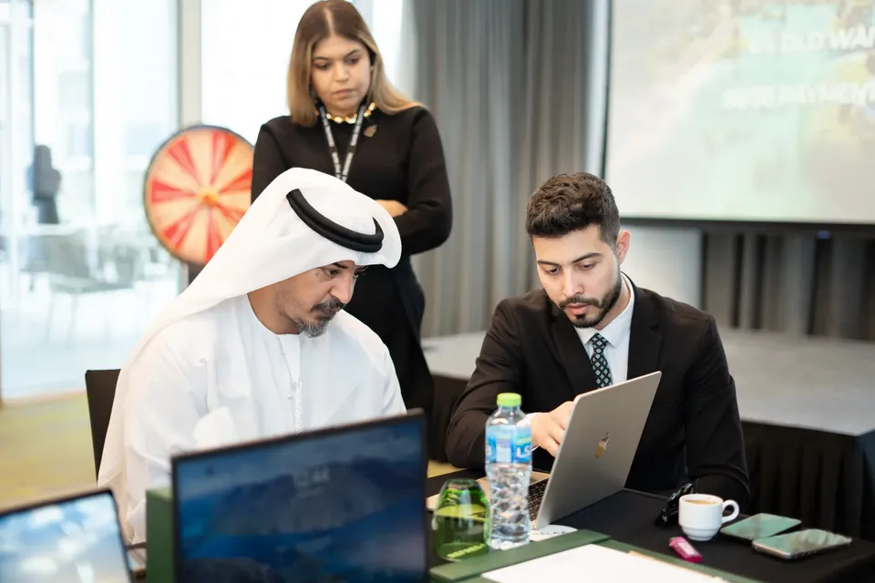 A visitor in a white kandura studies a laptop with an adviser, a colleague standing behind and a prize wheel in the background