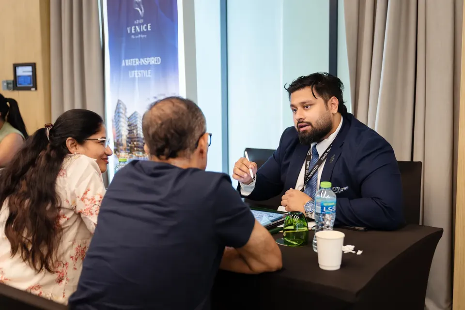 Gesturing as he talks, an adviser sits with two visitors at a table beside a project display banner