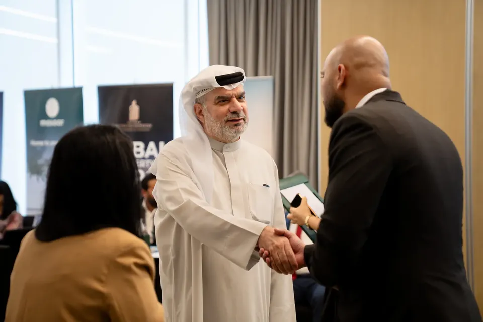 A man in a white thobe shakes hands with a guest in a dark suit, a woman in a mustard blazer nearby