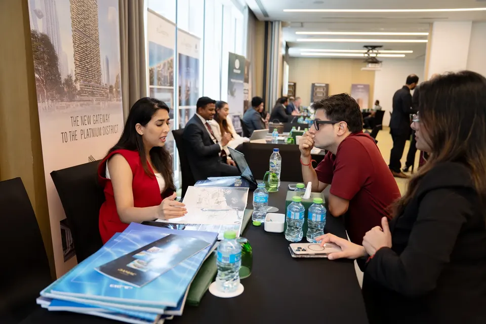A seated guest reviews brochures with an adviser in red, beside a project banner