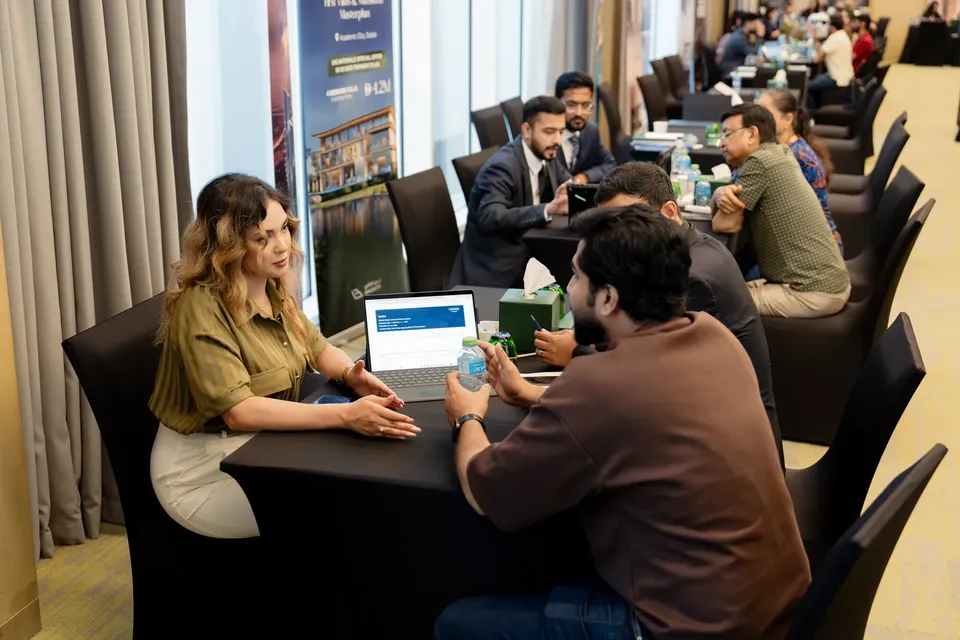 A woman consults a visitor over a laptop at a table, with tissues and water bottles between them, at the property expo