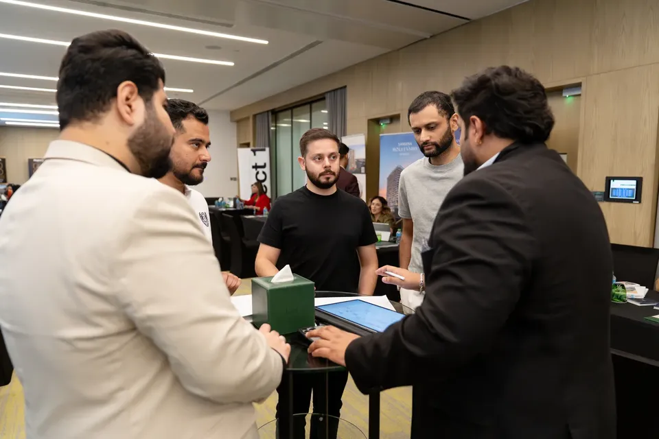 Five men gather around a small tablet display table at the expo, one in a dark suit gesturing