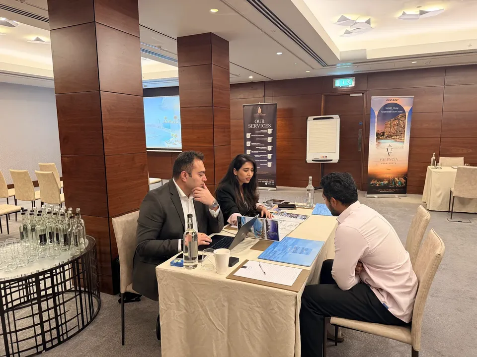 Two advisers consult with a visitor at a table, beside a services banner and a DAMAC Valencia display