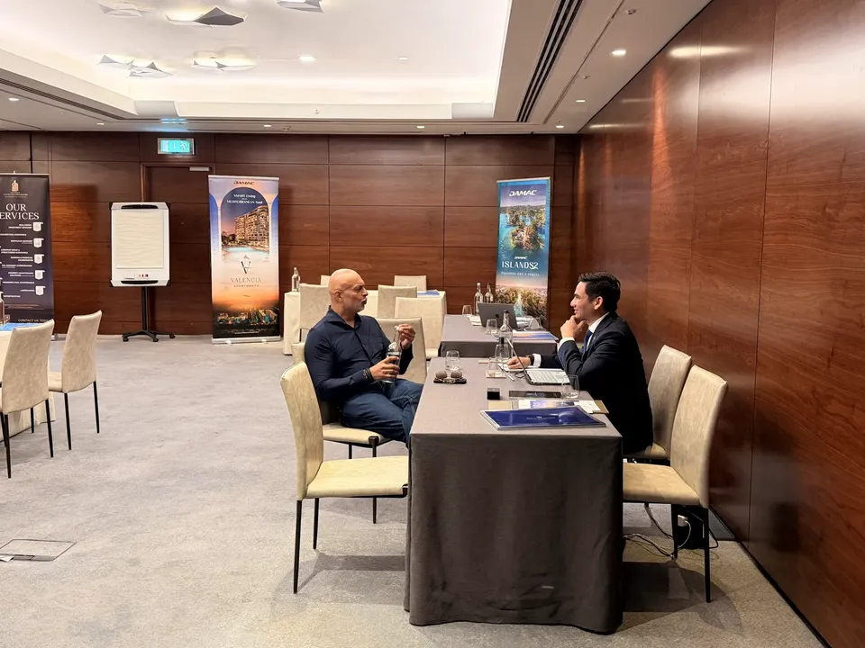 Two men talk at a table in front of DAMAC Valencia and Islands 2 project banners at the expo