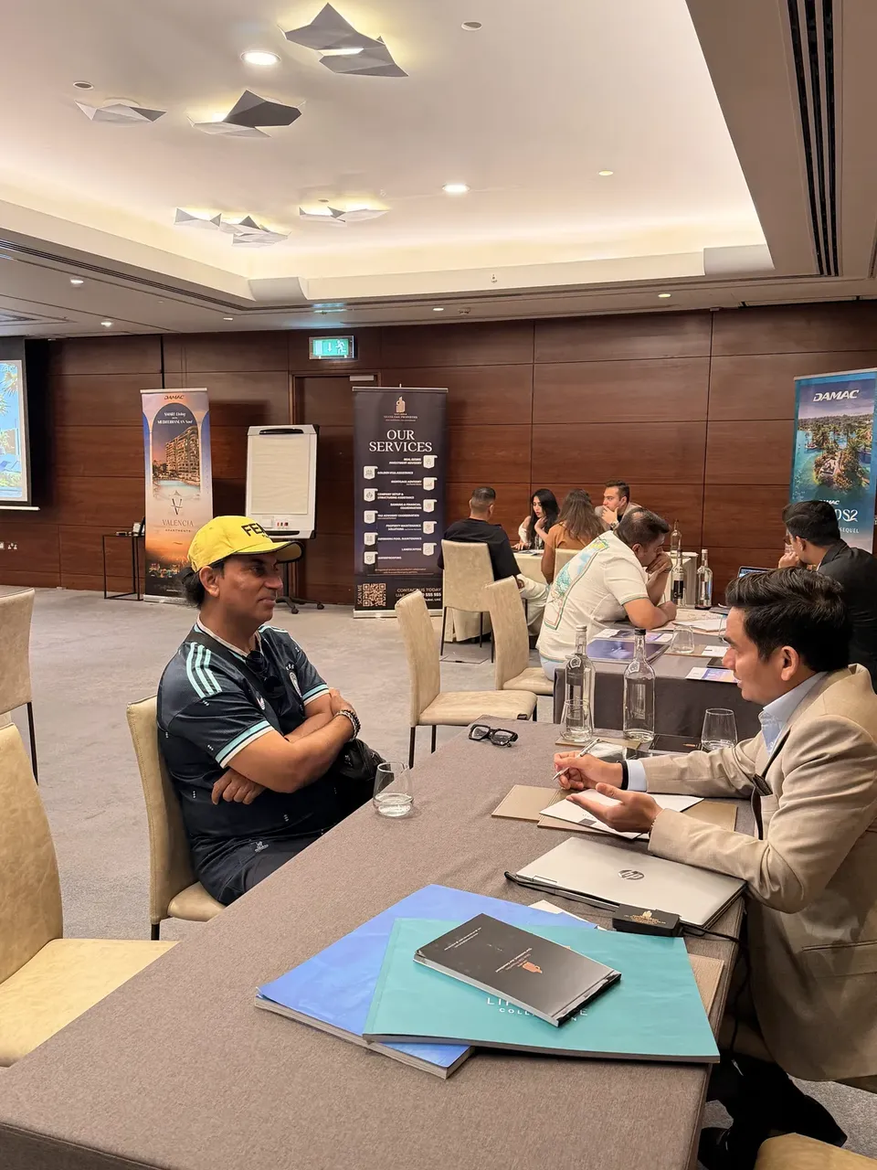 Wearing a yellow cap, a visitor sits across from an adviser, with other consultations continuing beside DAMAC project banners