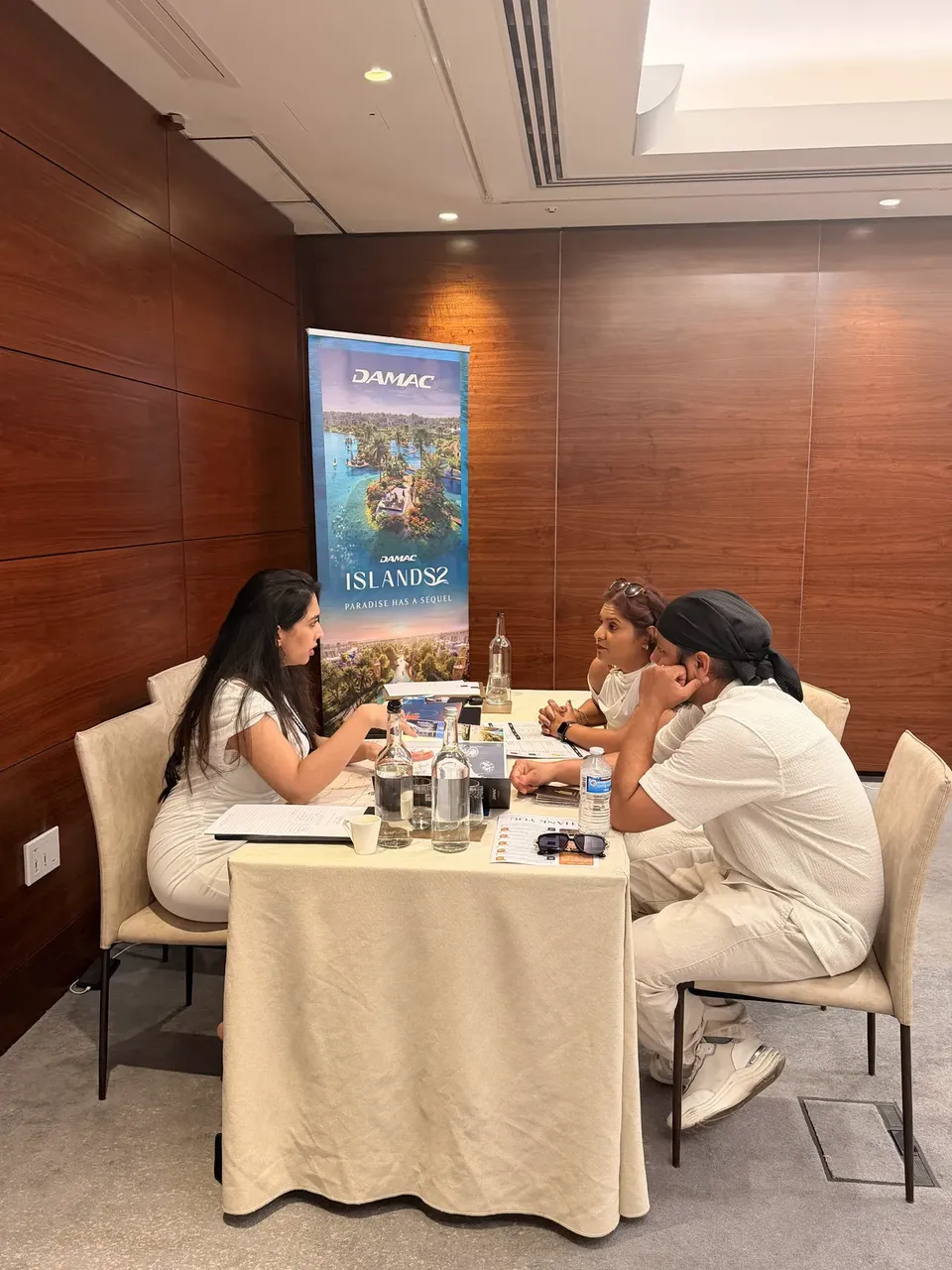 An adviser speaks with two visitors at a round table in front of a DAMAC Islands 2 display