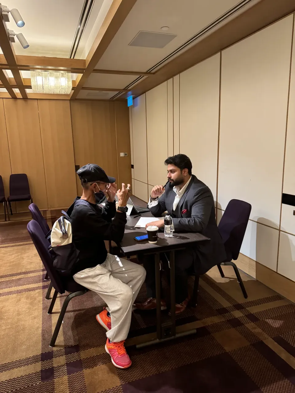 Two men sit across a small table in conversation at the expo, in a wood-panelled room with purple upholstered chairs