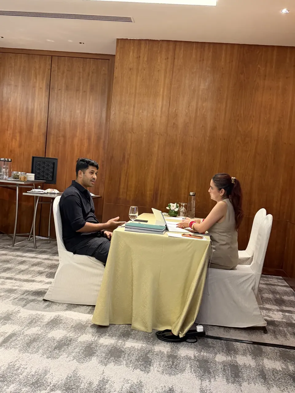 A man in a black shirt talks with a woman at a small round table covered with papers and folders