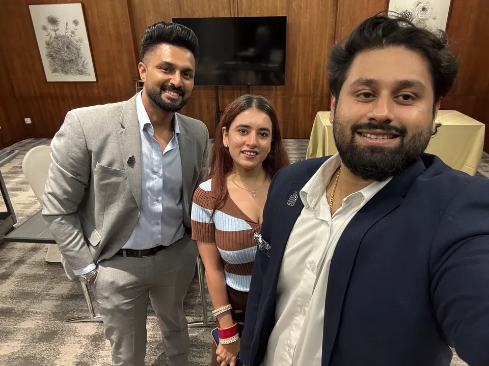 Three people smile for a selfie in a wood-panelled room, standing in front of a blank television screen and framed artwork