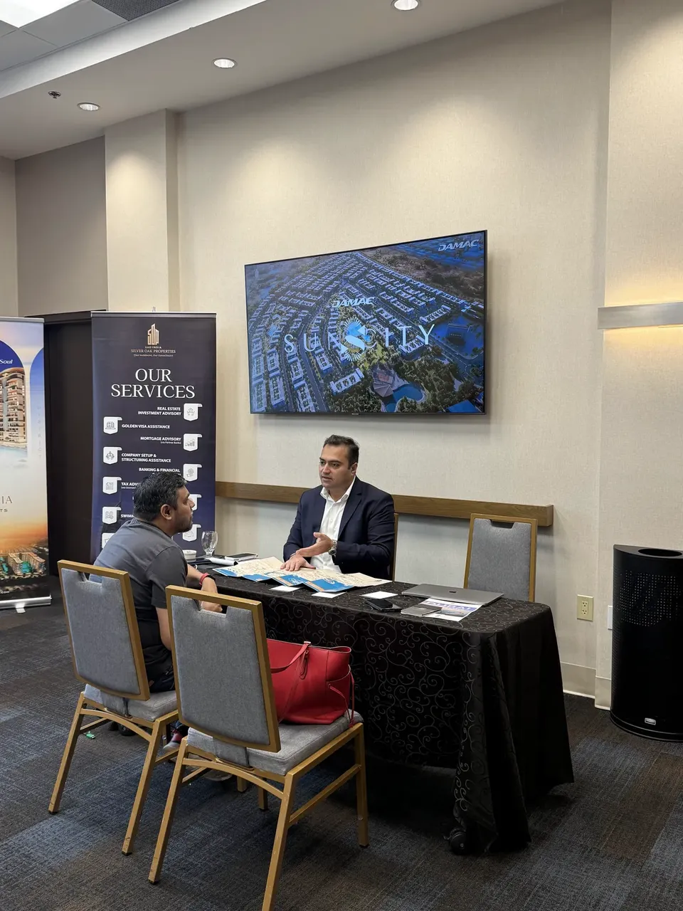 Two men sit at a table below a screen showing a DAMAC development, with a red bag on a nearby chair