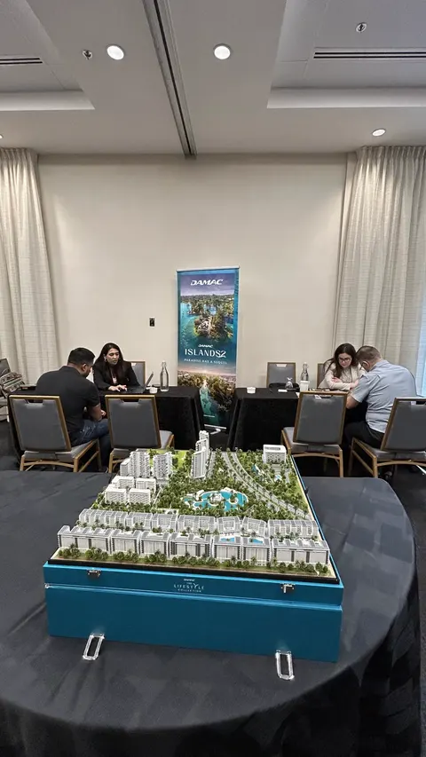 A scale model of a development sits in a blue display case near tables and a DAMAC Islands 2 banner