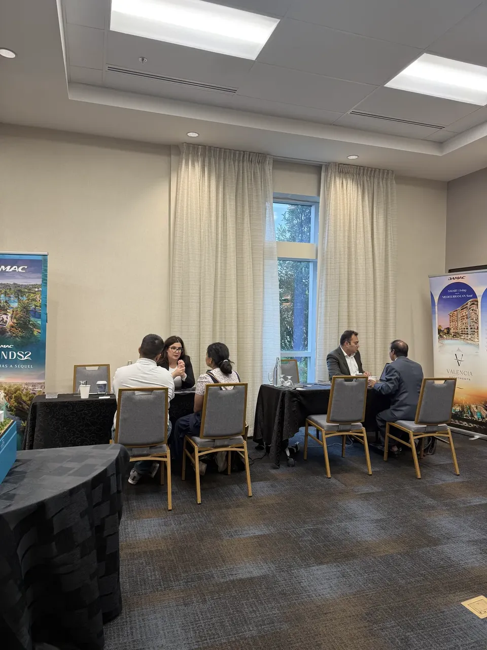 Two groups hold consultations at tables by a curtained window, with DAMAC and Valencia project banners along the walls