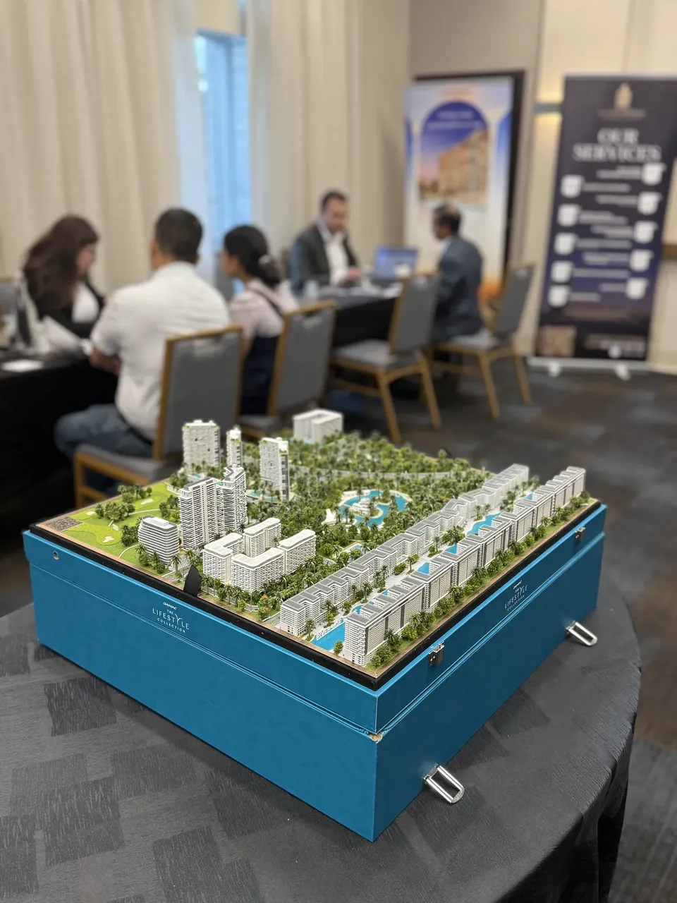 A scale model of a residential development displayed in an open blue presentation case, with a consultation underway behind