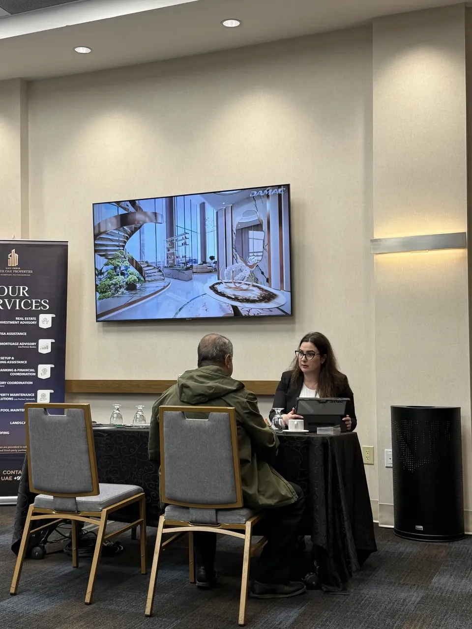 An adviser talks with a visitor at a small table at the expo, below a screen showing a DAMAC interior render