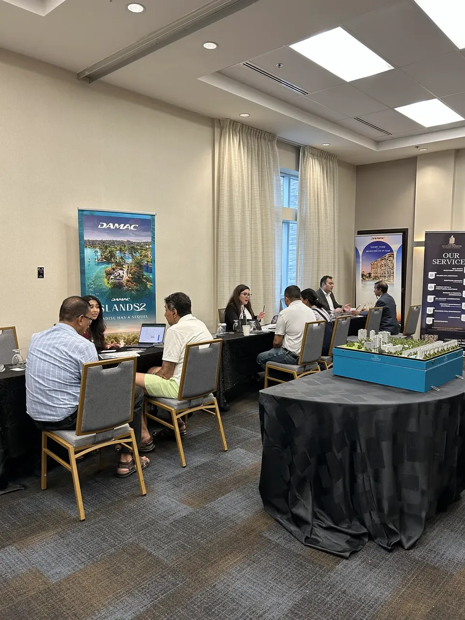 Several consultations take place along a shared table at the expo, in front of a DAMAC Islands 2 display banner